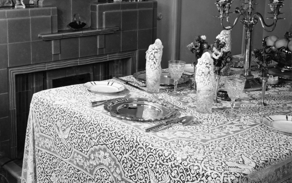 A black & white photo of an intricate lace tablecloth in Arts & Crafts style covering a dining room. The table also features silver tableware and cutlery, crystal glassware and a prominent candlestick.