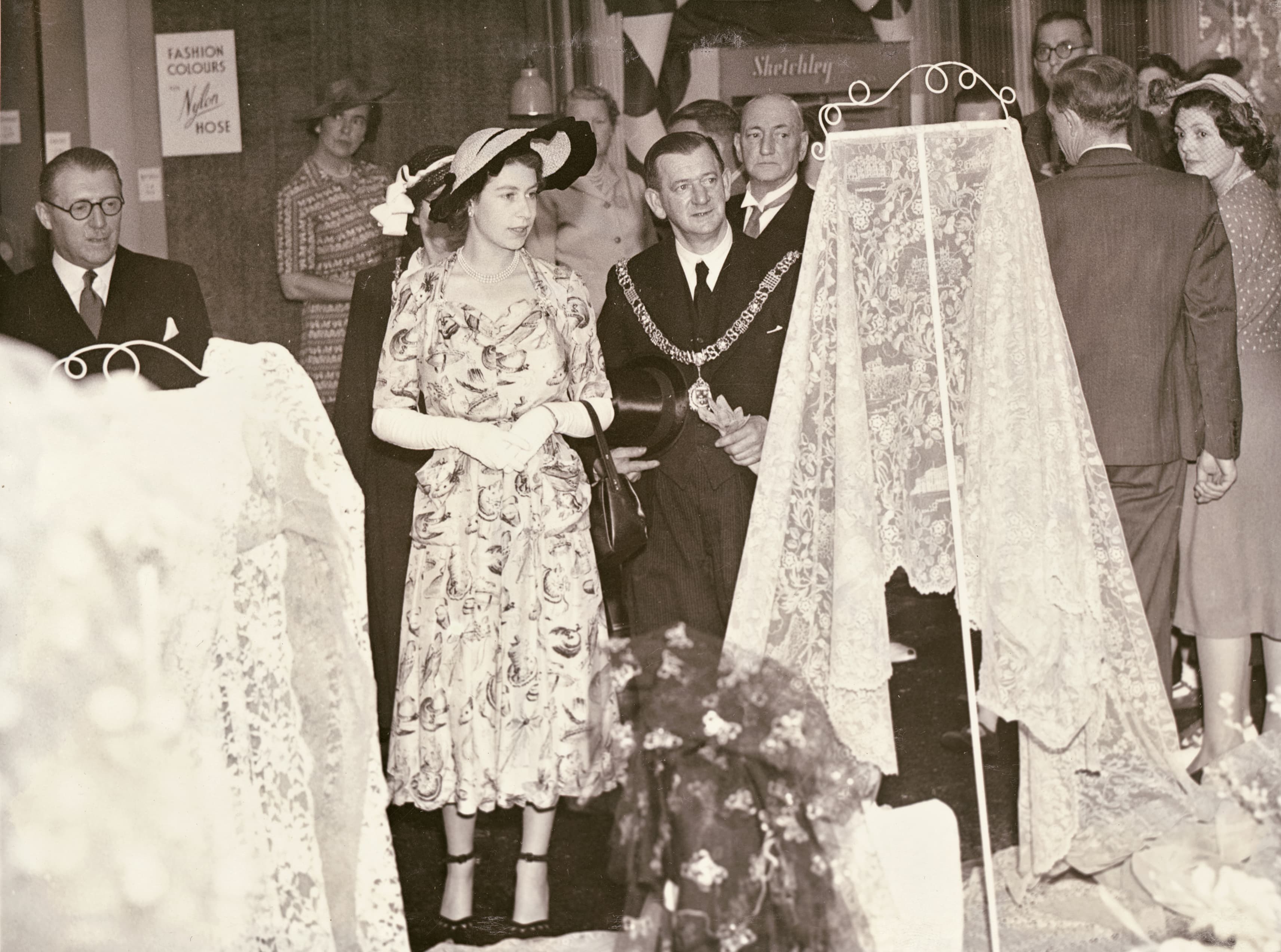 A black & white photo of Princess Elizabeth admiring some Nottingham lace during a 1949 visit to Nottingham. Local dignitaries look on beside her.