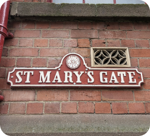 A red street sign made from iron with white text reading St Mary's Gate. There is a lace motif above the text.