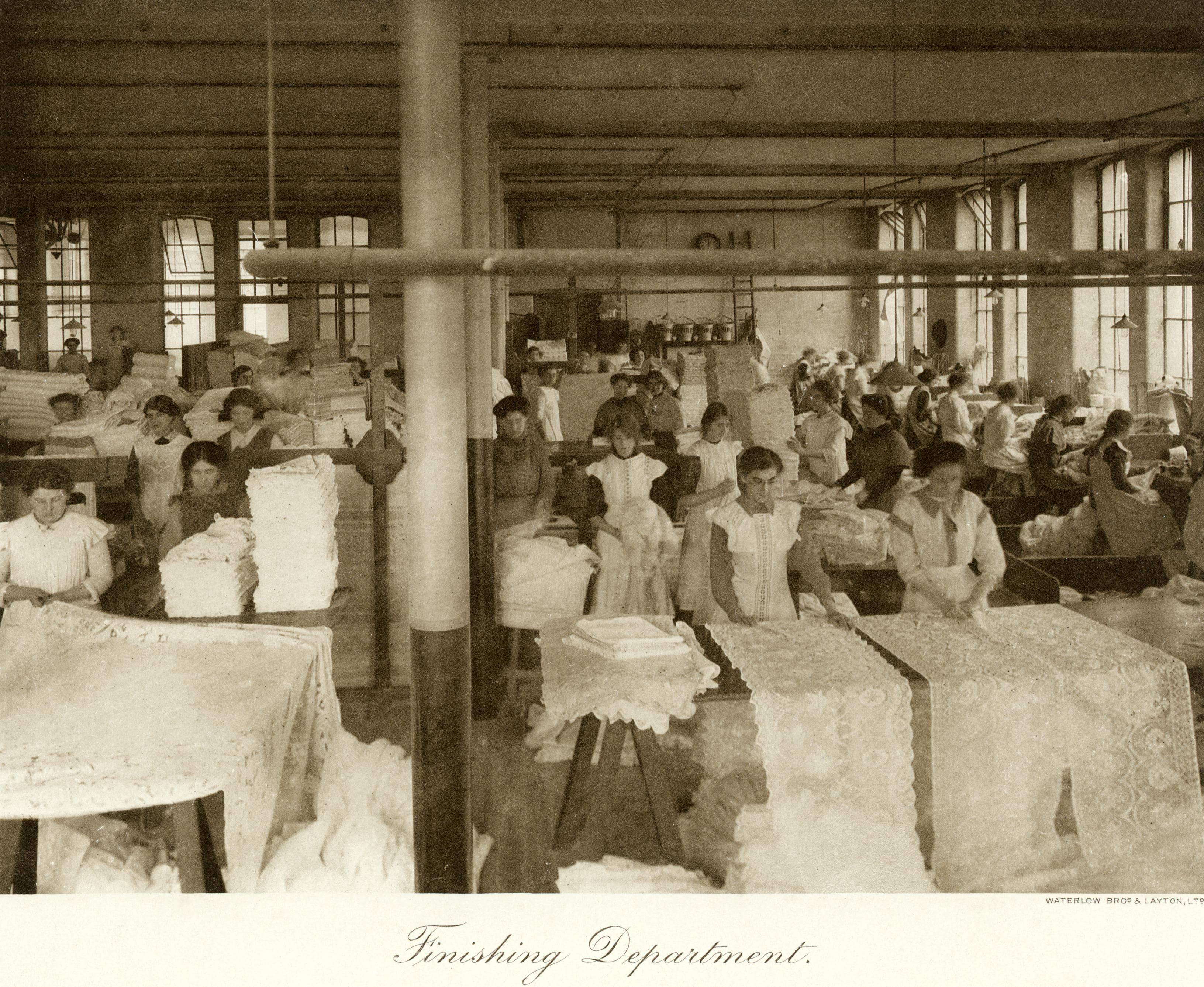 A warehouse room full of women work on finishing lace garments and drapery. At the bottom of the photo are the words 'Finishing Department'.