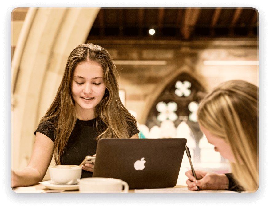 A young female student with a laptop at the library. She is smiling and has a cup of coffee next to her